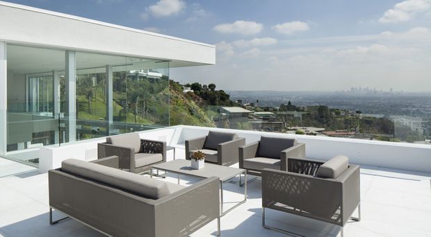 A bright modern rooftop terrace or balcony with a white rendered flat-roof structure, expansive floor-to-ceiling glazing, and a coordinated outdoor seating arrangement overlooking a sprawling urban landscape. The terrace uses pale hard surfaces, low white parapet walls, and grey contemporary lounge furniture under a clear blue sky.