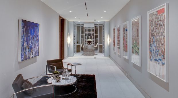 A long, refined interior gallery hallway with pale grey walls, large-format light flooring, recessed ceiling lighting, and a series of framed artworks displayed along both sides. A small seating area with sculptural chairs, a round glass coffee table, and a dark rug occupies the foreground, while a decorative screened opening and console area terminate the view.