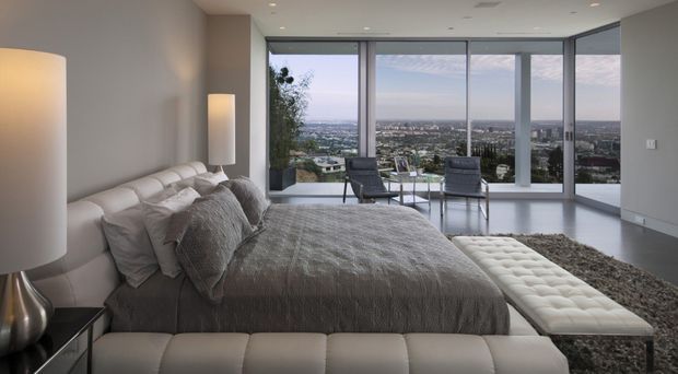 A sophisticated contemporary bedroom with a large upholstered bed, layered grey bedding, a tufted bench at the foot, and matching bedside lamps. Full-height corner glazing provides expansive city views, while a pair of lounge chairs sits beside the windows. The room has a neutral grey and white palette, polished minimalist detailing, and abundant natural light.