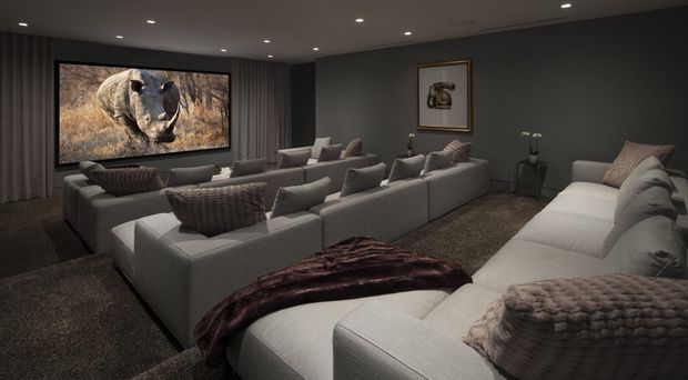 A dark, luxurious home cinema with multiple large light-grey sectional sofas arranged toward a wall-mounted projection screen displaying wildlife imagery. The room has charcoal walls, full-height dark curtains, recessed ceiling spotlights, deep textured carpeting, and plush patterned cushions and throws. Framed artwork and a small side table with an orchid add refined decorative details.