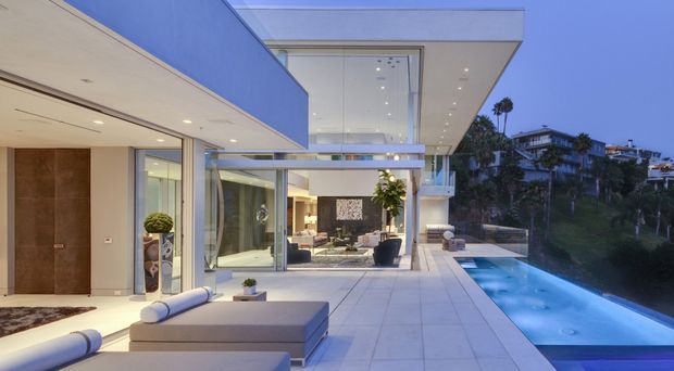 Contemporary multi-level luxury residence viewed from an open terrace at dusk, with expansive floor-to-ceiling glazing, crisp white rendered planes, a large cantilevered upper volume, and a long narrow swimming pool along the edge. The terrace and interior lounge are visible, with landscaped hillside residences in the background.