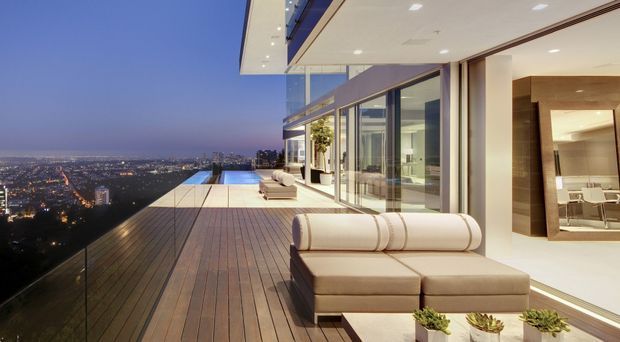 A luxurious high-rise residence with a large timber-decked terrace overlooking a city at dusk. The image shows floor-to-ceiling glazing, a deep roof overhang with recessed lighting, streamlined outdoor lounge seating, glass balustrades, and a warm contemporary interior visible through the open glazing. The terrace includes a long narrow swimming pool or reflective water feature along the edge.
