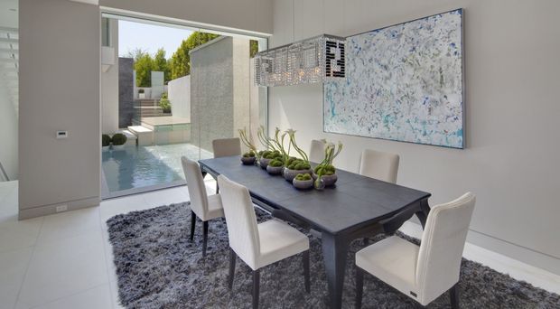 Bright contemporary dining room with a dark rectangular dining table, six upholstered dining chairs, and sculptural pendant lighting. A large abstract artwork hangs on a pale wall, while floor-to-ceiling glazing opens the room toward a narrow water feature and landscaped courtyard. The space has a restrained neutral palette with white tiled flooring and a textured grey rug.