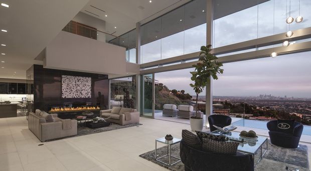A luxurious contemporary double-height living room with expansive floor-to-ceiling glazing overlooking a cityscape. The open-plan space features a suspended fireplace wall, large pale tile flooring, sculptural contemporary furniture, a glass coffee table, a mezzanine, and dramatic linear pendant lighting. An outdoor terrace and pool are visible beyond the glazing.