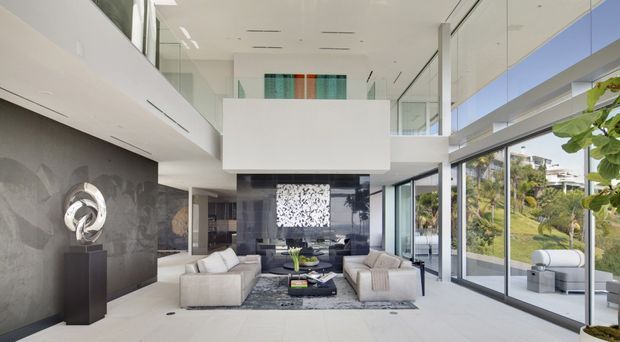 A bright, sophisticated double-height living room with floor-to-ceiling glazing, a large suspended white architectural volume, and a mezzanine-level glass balustrade. Pale sofas face a dark feature wall and fireplace/media area, while a sculptural artwork stands on a dark plinth. The room opens visually to landscaped grounds and has a clean, contemporary character.