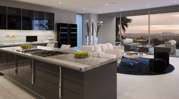 A spacious open-plan contemporary kitchen and living area with a large dark built-in island, pale stone benchtops, integrated appliances, glossy dark cabinetry, and a white tiled floor. Floor-to-ceiling glazing opens to a panoramic urban view. The adjoining living zone contains light sofas, a dark rug, coffee tables, and sculptural seating, creating a polished high-rise interior.