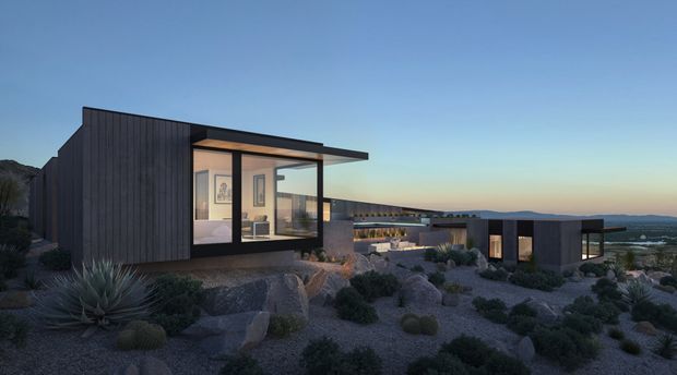 A contemporary low-rise residential complex set in a rugged, sparsely planted landscape at dusk. The buildings feature dark vertical cladding, expansive floor-to-ceiling glazing, strong horizontal roof planes and flat-roofed forms, with warm interior lighting visible through the windows. Native-style drought-tolerant planting, rocks and gravel surround the structures, with distant open terrain and water visible beyond.