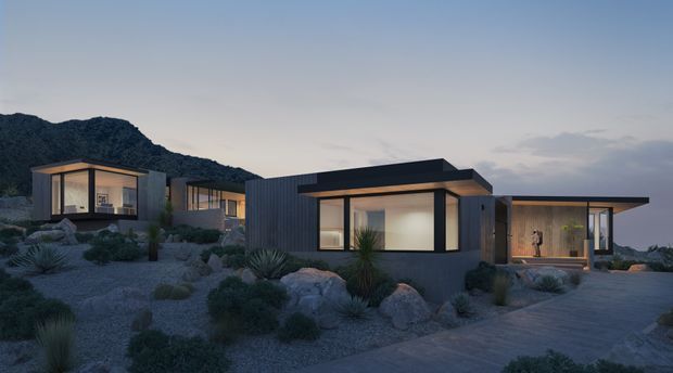 A low-profile modern residential compound composed of several connected flat-roofed volumes, set within a rocky, drought-tolerant landscaped site. The dark vertical cladding and expansive glazed openings are softly illuminated at dusk, with rugged mountains forming the background.