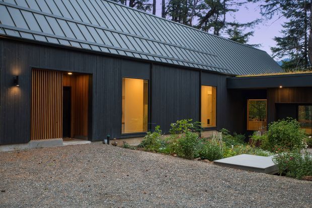 Exterior view of a dark, contemporary single-storey building with a long standing-seam metal pitched roof and vertically textured dark cladding. Warm light glows through tall windows and an entry door, while a gravel forecourt and planted garden border the facade.