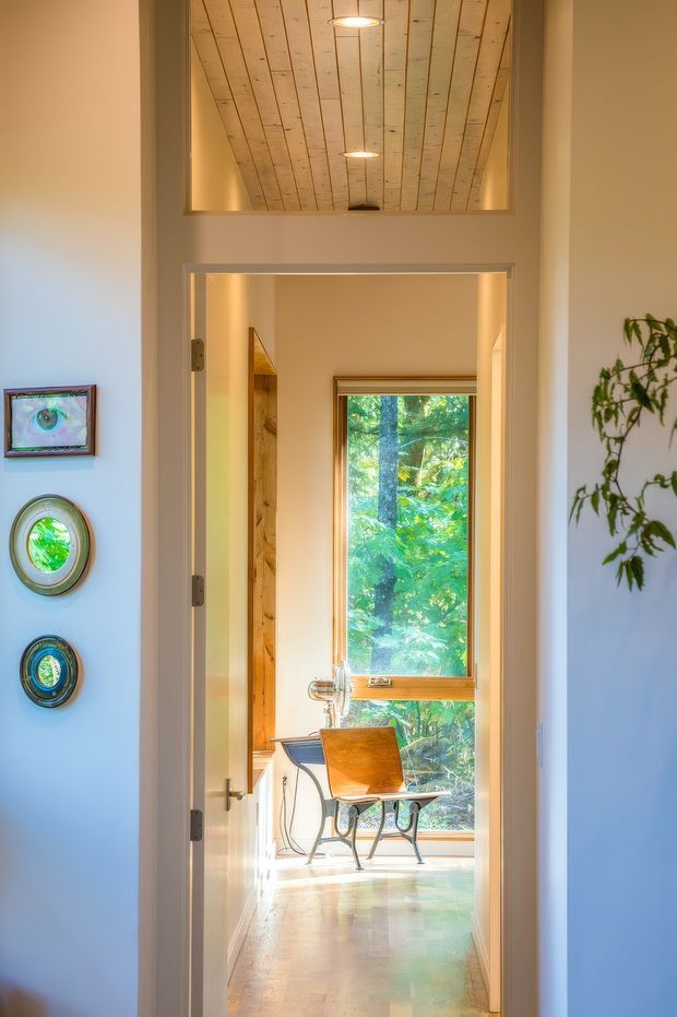 A bright interior view through an open doorway into a compact home office or study. The room beyond has a small desk, a wooden chair, a tall timber-framed window overlooking greenery, and warm natural light. A distinctive plank-lined timber ceiling is visible above the doorway, while pale painted walls, timber trim, polished flooring, framed artwork, and a potted plant create a warm, eclectic atmosphere.