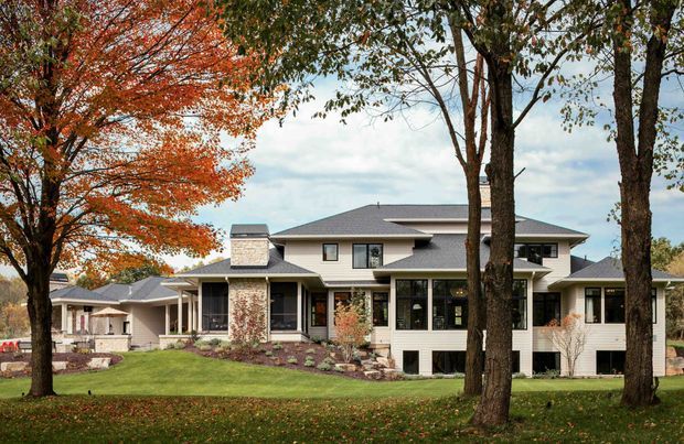 A large suburban two-storey residence viewed from a landscaped front lawn, with a broad low-pitched roof, multiple gabled and hipped roof sections, light horizontal siding, stone-clad chimney and porch columns, dark-framed windows, and extensive mature deciduous trees in an autumn setting.