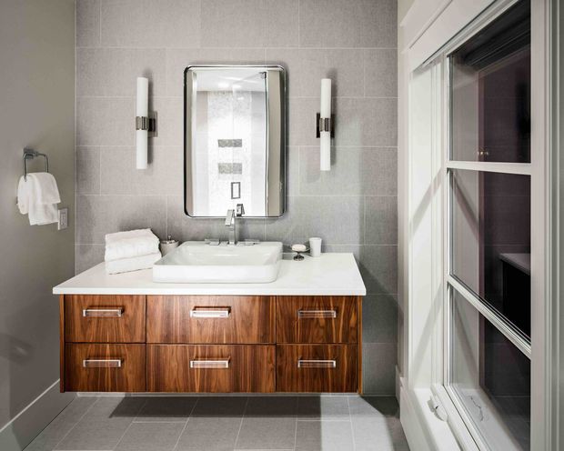 A compact, refined bathroom centered on a floating timber vanity with a white vessel basin and white benchtop. The walls and floor are finished in large-format grey tiles, with a rectangular framed mirror, paired wall sconces, chrome tapware, and a large white-framed window providing natural light.