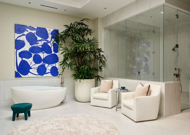 Bright, refined contemporary bathroom with a freestanding white bathtub, large frameless glass shower enclosure, pale stone or tile surfaces, sculptural white lounge chairs, a small teal stool, a large leafy plant, and bold blue abstract wall art. The room has a calm, luxurious spa-like atmosphere with recessed ceiling lighting and extensive mirrored or reflective glazing around the shower.