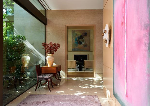 A refined interior passage or entry hall with full-height glazing along one side, a pale stone or plaster feature wall, timber wall panelling, and a large pink-toned artwork. A small dark timber console with a vase and chair sits beside the glazing, while a framed opening leads to a sitting room with a fireplace and artwork. Dappled natural light creates a calm, gallery-like atmosphere.