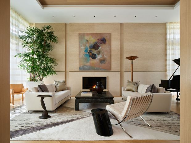 A spacious, refined living room with a symmetrical seating arrangement around a dark stone coffee table and a linear fireplace. The room features warm beige stone or plaster wall panels, a timber-lined recessed ceiling, floor-to-ceiling sheer curtains, large pale patterned rugs, sculptural lounge chairs, a grand piano, indoor greenery, and understated contemporary furnishings. An abstract artwork hangs above the fireplace, while a lit fire adds warmth and ambience.