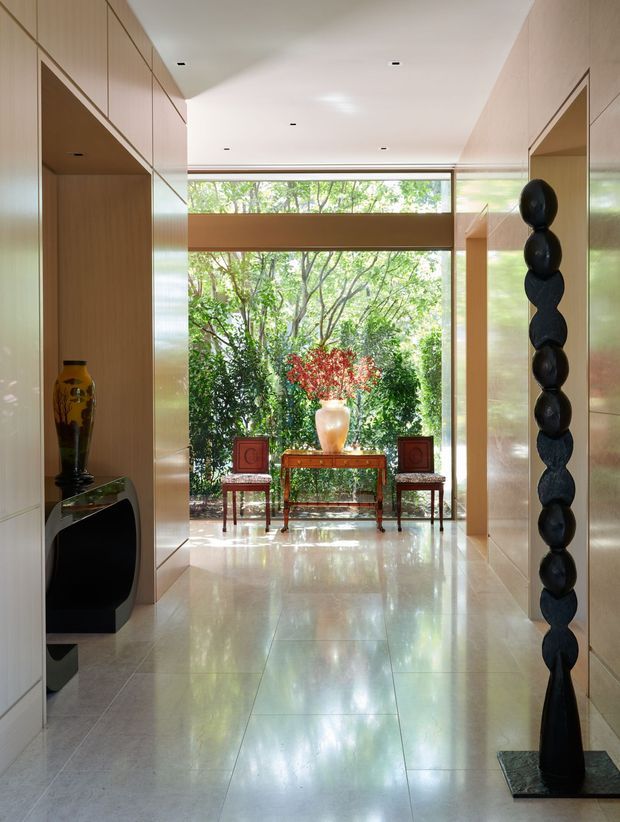 A bright, sophisticated interior entry hall with floor-to-ceiling glazing at the far end overlooking lush greenery. Pale timber wall panelling and built-in display niches frame a polished light stone-tile floor, while curated sculptural objects, a console table, chairs, and a large vase create a refined gallery-like composition.