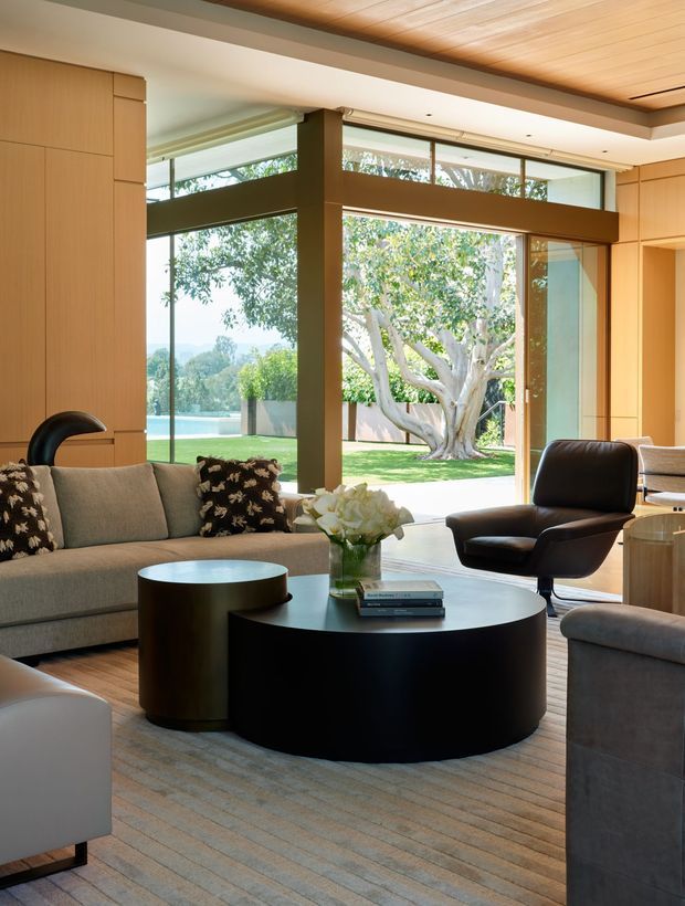 A refined living room with extensive floor-to-ceiling glazing overlooking a landscaped garden and pool. The space features warm wood wall panelling and a wood-slat ceiling, dark structural window framing, a large neutral sectional sofa, sculptural black coffee tables, and a black leather lounge chair. Natural daylight creates a calm, sophisticated atmosphere.