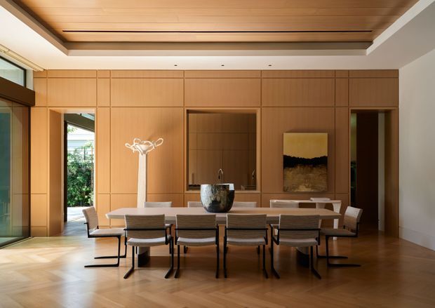A refined dining room with a long rectangular dining table and multiple upholstered chairs, set against a full-height warm timber-paneled wall. The room features integrated cabinetry and openings, a recessed timber-lined ceiling with concealed lighting, timber flooring, expansive glazing, and restrained contemporary furnishings and artwork.
