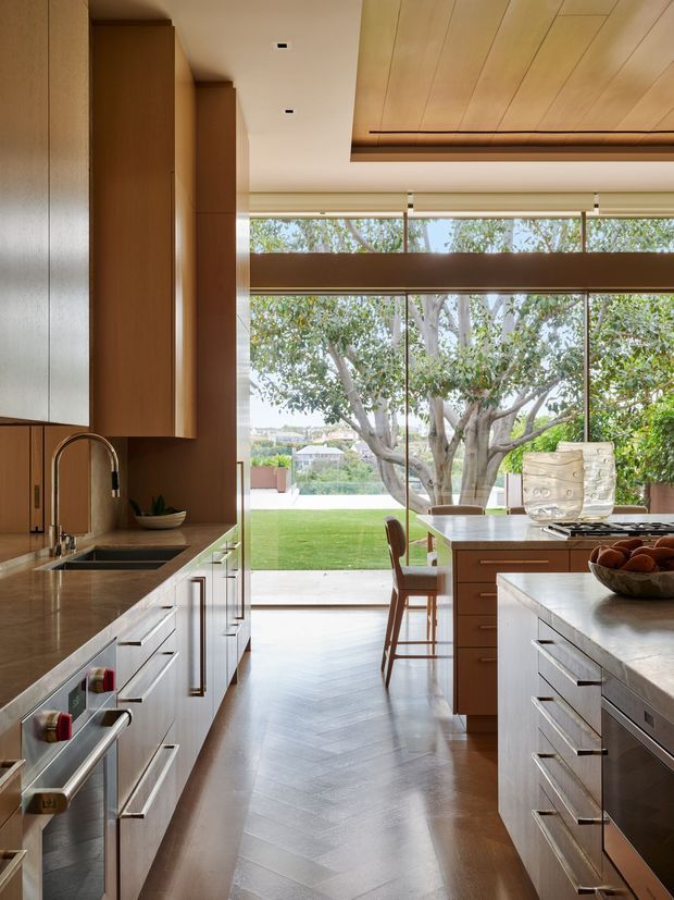 A refined, spacious contemporary kitchen viewed along a central aisle toward a dining area and expansive garden-facing glazing. The kitchen features extensive warm timber cabinetry, stone benchtops, integrated stainless-steel appliances, a large island, a timber-lined ceiling recess, and herringbone timber flooring. Natural light, mature trees, and a lawn are visible beyond the glass doors.