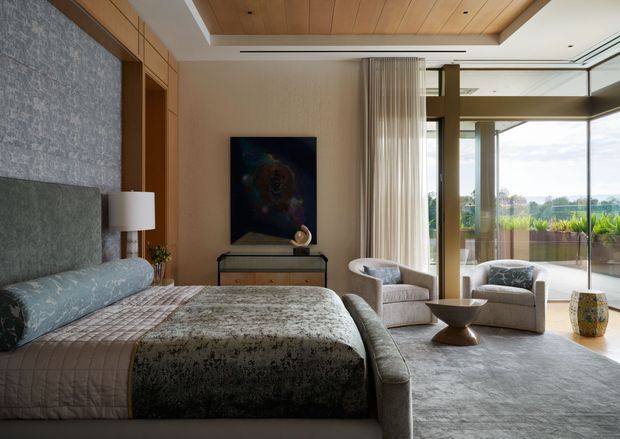 A sophisticated bedroom with a large upholstered bed, textured grey-blue feature wall, and warm timber-lined recessed ceiling. The room includes a compact dark timber console, bedside lighting, two upholstered lounge chairs, a sculptural side table, and full-height sheer curtains opening to expansive glazing and a balcony or terrace with planted screening. The palette is muted and refined, combining grey, taupe, cream, and warm timber tones.