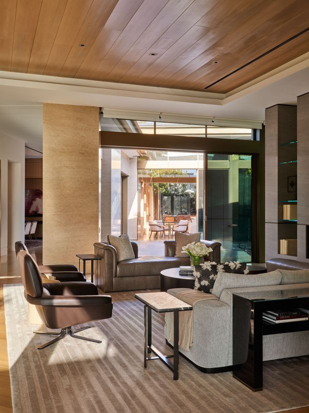 A refined contemporary living room with a warm timber-panelled recessed ceiling, pale stone feature wall, dark built-in shelving, and expansive glazed doors opening to a bright courtyard or outdoor dining area. The room is furnished with a large sectional sofa, leather lounge chairs, nesting side tables, a round coffee table, and a large neutral striped rug. Natural light, layered textures, and restrained earthy tones create an elegant, inviting atmosphere.