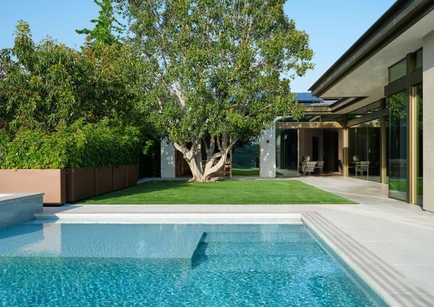 A contemporary single-level home opening onto a landscaped courtyard and swimming pool. The image shows broad floor-to-ceiling glazing, dark slim window frames, pale rendered walls, a projecting flat roof, and a light paved terrace surrounding a bright blue pool. Mature trees and hedging provide a lush private setting, while the open-plan interior is visible through the glazing.