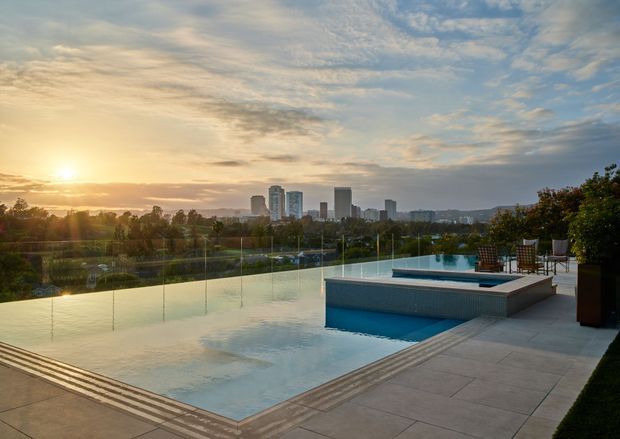A rooftop or elevated outdoor terrace with a large infinity-edge swimming pool overlooking a landscaped park and distant urban skyline at sunset. The pool is bordered by pale stone-look paving, a glass balustrade, outdoor dining furniture, and large planters. The scene has expansive views, warm sunset light, and a refined contemporary resort-like atmosphere.