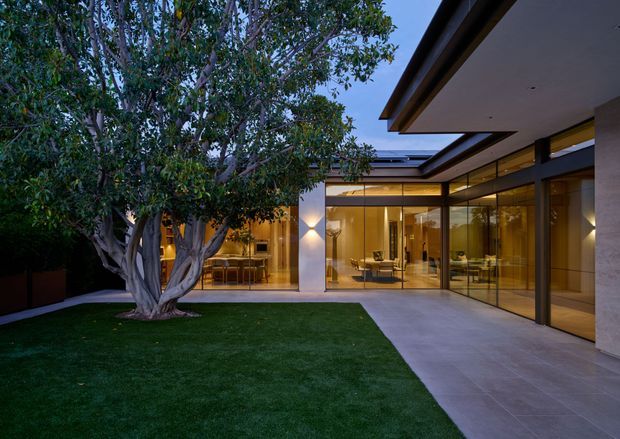 Twilight view of a refined contemporary residence arranged around a manicured courtyard. Floor-to-ceiling glazed walls open the interior toward a grass lawn and mature sculptural tree, while broad roof overhangs, dark metal window frames, pale rendered walls, and warm interior lighting create a sheltered indoor-outdoor connection.