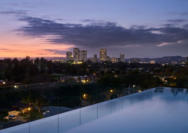 Twilight cityscape viewed from an elevated rooftop or terrace, with a reflective infinity pool in the foreground behind clear glass balustrades. A suburban neighbourhood, illuminated high-rise buildings, and distant hills sit beneath a dramatic purple and orange sunset sky.