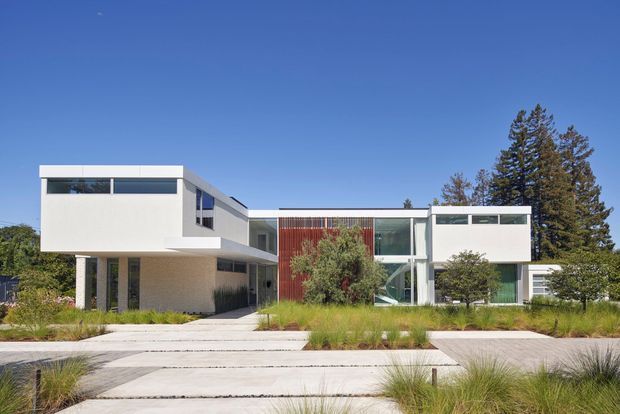 Front exterior view of a contemporary multi-wing residence with crisp white cubic forms, extensive horizontal glazing, a projecting upper volume supported by slender columns, and a vertical red-toned feature cladding panel. The house is set behind a landscaped front garden with ornamental grasses, paved pedestrian paths, and mature trees under a clear blue sky.