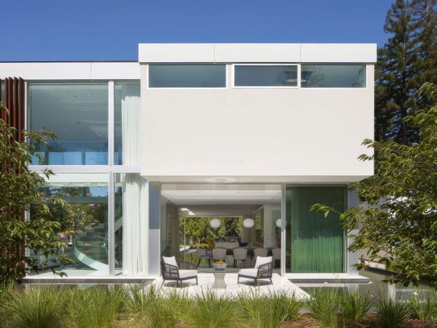 Front exterior view of a modern two-storey residence with a crisp white rectilinear facade, extensive floor-to-ceiling glazing, projecting upper-level volume, and a sheltered outdoor entertaining area opening toward landscaped planting. The architecture is clean-lined and minimalist, with a flat roof and strong indoor-outdoor connection.