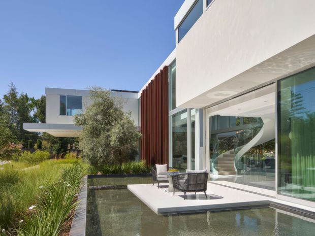 Exterior architectural view of a contemporary multi-level residence with crisp white rendered volumes, extensive floor-to-ceiling glazing, a vertical timber screen, and a landscaped courtyard. A shallow reflecting pool borders a white paved terrace with outdoor lounge chairs, while a sculptural curved staircase is visible through the glass.