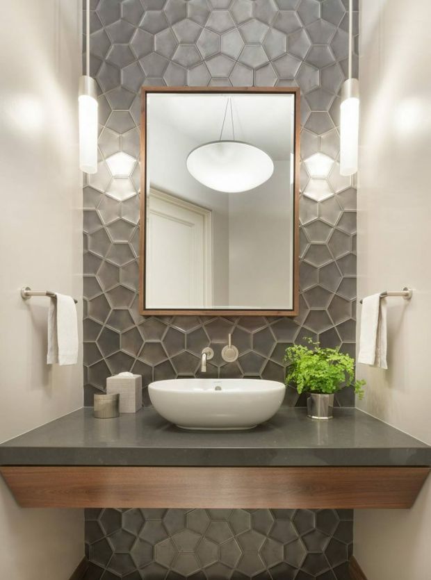 A compact, refined bathroom vanity with a floating timber-front cabinet, dark stone benchtop, white vessel basin, and brushed metal wall-mounted tapware. The feature wall is clad in dimensional grey hexagonal tiles, framed by pale painted walls and illuminated by slim vertical wall sconces. A timber-framed rectangular mirror reflects a suspended round pendant light, while towels and a small potted plant add functional and natural accents.