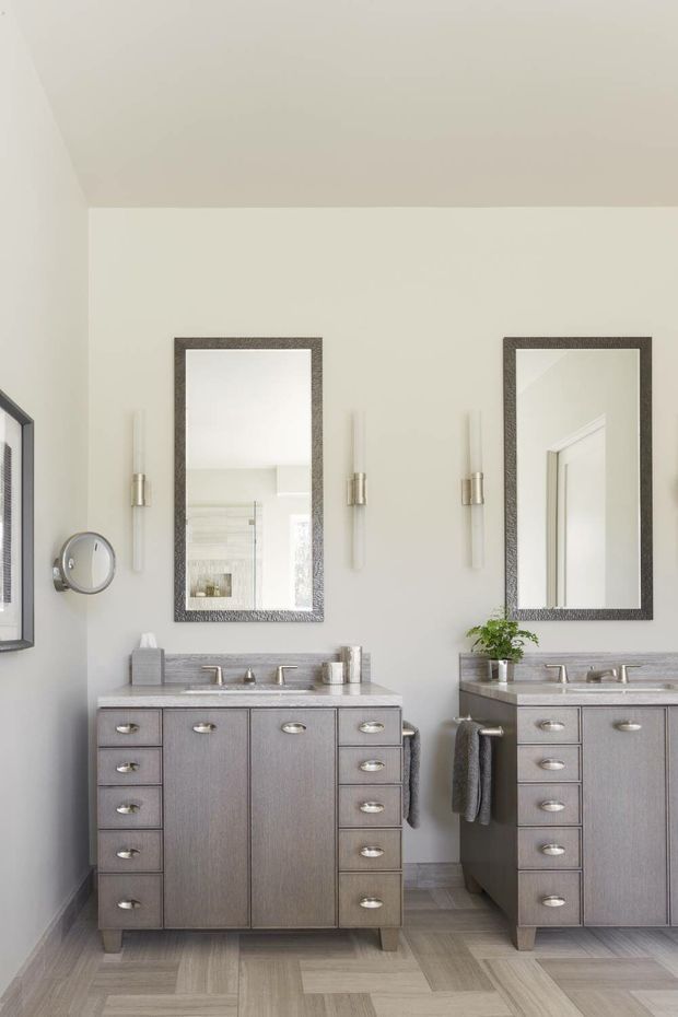 Bright, refined bathroom featuring two symmetrical grey wood-tone vanity units with integrated stone-look benchtops, undermount basins, chrome fixtures, framed mirrors, wall sconces, and tiled flooring. The space has a calm neutral palette and abundant natural light.