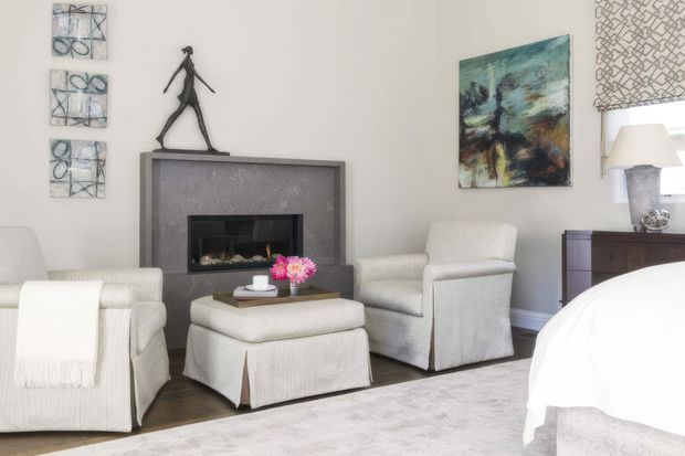 Bright, refined bedroom sitting area with a fireplace, two upholstered armchairs, a matching ottoman, abstract artwork, and a dark wood console. A partially visible bed and bedside lamp establish the room as a bedroom. The palette is predominantly soft white, cream, light grey, and dark wood, with restrained contemporary-classic styling.