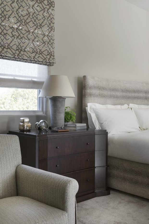 A refined bedroom corner featuring a neatly made upholstered bed with a tall textured grey headboard, a dark wood bedside chest, and a light upholstered armchair in the foreground. A layered window treatment combines a patterned Roman blind with a sheer roller shade, while a sculptural table lamp, books, metallic accessories, and a small plant add detail. The room uses a soft neutral palette with tailored, traditional furnishings and plush carpeting.