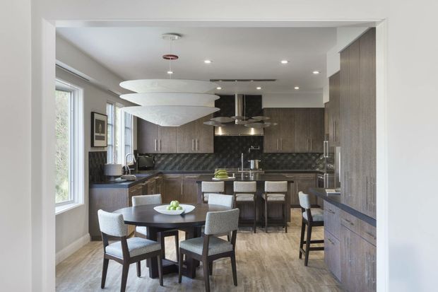 A contemporary open-plan kitchen and dining area viewed through a wide doorway. The kitchen features dark wood-toned cabinetry, a patterned dark backsplash, a central island with bar seating, stainless-steel appliances and fittings, and pale timber-look flooring. A round dining table with upholstered chairs sits in the foreground, illuminated by sculptural white pendant lights and recessed ceiling lighting. Large windows provide natural light.