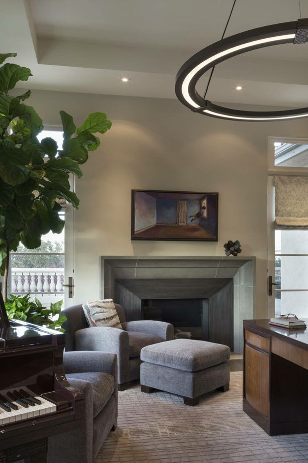 A refined living room with a grey upholstered armchair and ottoman arranged around a substantial grey stone or tiled fireplace. A grand piano occupies the foreground, with a timber sideboard at right, framed artwork above the mantel, indoor greenery, glazed French doors, and a large contemporary circular pendant light. The room has a warm neutral palette and a formal yet comfortable character.