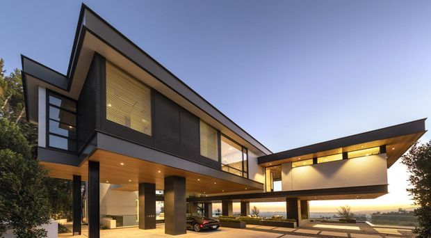 Exterior view of a large contemporary two-storey residence with sharply angled roof planes, dark cladding, extensive glazing, warm timber soffits, and a partially open elevated ground level. The house includes a covered carport/drive-through area, landscaped foreground, and broad distant views beyond the site.