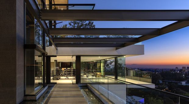 A dusk view from a contemporary multi-level residence, looking along a glazed terrace or walkway beneath a strong exposed steel pergola structure. Floor-to-ceiling glass walls and balustrades open toward a distant urban or coastal horizon, while warm interior lighting reveals a refined living or entertaining area inside. The composition emphasizes cantilevered horizontal planes, transparent glazing, and the contrast between cool twilight and warm illumination.