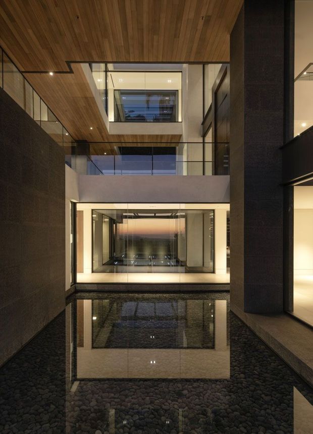 A dramatic multi-level interior courtyard or atrium with a shallow reflective water feature over a bed of dark rounded stones. Floor-to-ceiling glazing, dark textured wall surfaces, warm timber ceiling lining, exposed concrete, and integrated recessed lighting frame the tall void. The space has a refined, minimalist contemporary character with strong vertical layering and reflections.