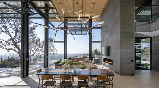A sophisticated open-plan dining and living area with floor-to-ceiling glazing overlooking a city and water view. The room features a long timber dining table with contemporary wooden chairs, a light-toned sectional sofa, a linear wall-mounted fireplace set into a dark textured feature wall, expansive dark-framed windows, a timber-lined ceiling, and a sculptural modern pendant light. A stairway and additional interior space are visible to the right.