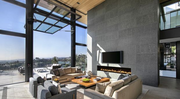 A bright, spacious contemporary living room with floor-to-ceiling black-framed glazing opening to a terrace and expansive views. The room features a large grey textured feature wall with a wall-mounted television and linear built-in fireplace, a timber-lined ceiling, neutral upholstered sofas and armchairs, and a substantial timber coffee table. A double-height glazed opening and upper-level interior balcony are visible to the right.