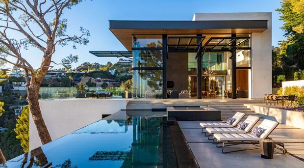 A modern multi-level residence viewed from its pool terrace, featuring a dramatic flat roof, floor-to-ceiling glazing, dark structural framing, warm timber-toned interior surfaces, and expansive outdoor entertaining areas. An infinity-edge pool and sun loungers occupy the foreground, with landscaped surroundings and a suburban hillside setting beyond.