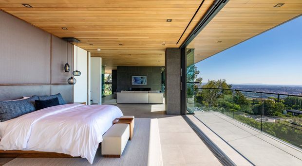 A contemporary bedroom with a low-profile bed, white bedding, upholstered bench, and dark blue cushions. The room opens to a broad glazed terrace or balcony with frameless glass balustrading and expansive coastal or urban views. A warm timber-lined ceiling with recessed lighting extends across the interior and outdoor edge, while dark stone or textured wall surfaces, integrated pendant lights, and a partially visible living area create a refined open-plan suite.