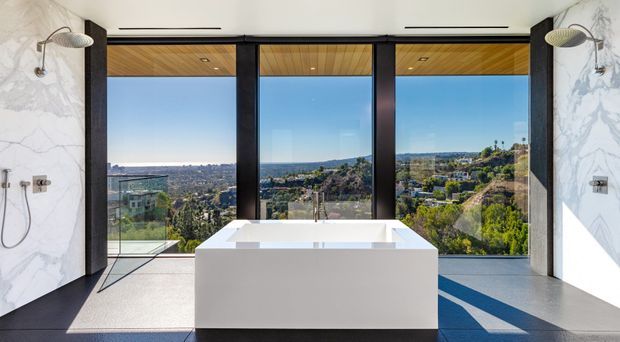 A luxurious contemporary bathroom with a freestanding white bathtub positioned in front of full-height black-framed glazing. The room features dramatic white marble-look wall surfaces, wall-mounted shower fixtures, a timber-lined ceiling, and expansive views over a hilly urban landscape. Strong natural light creates a refined, open atmosphere.
