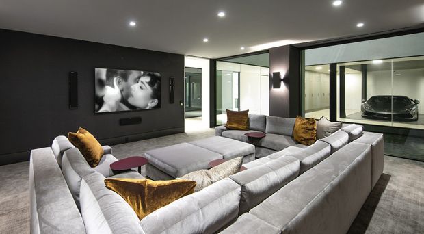 A sophisticated contemporary media room with a large U-shaped light-grey sectional sofa arranged around a low central coffee table and wall-mounted television. The dark feature wall, recessed ceiling lights, grey carpet, black-framed full-height glazing, and illuminated adjoining garage displaying a car create a luxurious, cinematic atmosphere.