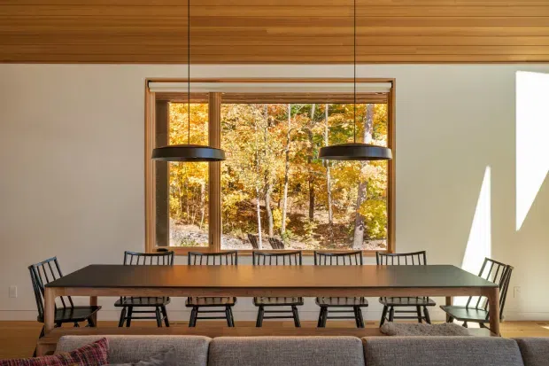 A warm, contemporary dining area with a long rectangular timber dining table and black spindle-back chairs, positioned in front of a large window overlooking an autumn woodland. Two black pendant lights hang above the table, while a timber-lined ceiling and pale walls create a clean, inviting interior. The back of a sofa is visible in the foreground.