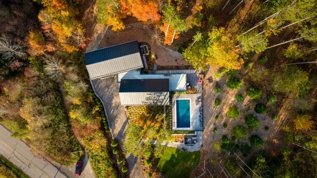 Aerial view of a contemporary residential property set within dense, mature woodland. The home consists of connected rectangular volumes with dark metal pitched roofs, surrounded by extensive trees, landscaped garden areas, paved paths, and a narrow rectangular swimming pool. A road and parked car are visible at the edge of the site.
