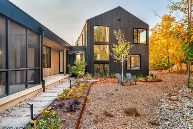 Exterior view of a contemporary multi-volume residence set within a wooded landscape. The building combines dark vertical timber cladding, expansive floor-to-ceiling glazing, warm timber soffits, and a simple gabled form. A gravel courtyard and stepping-stone path connect the wings, with restrained native-style planting and outdoor seating.